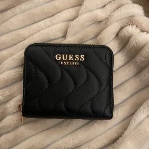 Svart quiltad plånbok från Guess - Snygg svart quiltad plånbok från Guess med guldfärgad dragkedja och logotyp. Perfekt för att hålla dina kort och kontanter organiserade. Stilren design som passar alla tillfällen.