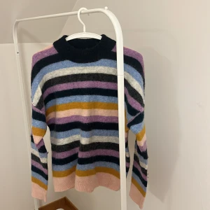 Randig stickad tröja från Second Female - Säljer en mysig stickad tröja från Second Female med färgglada ränder i rosa, blå, svart, vit och gul. Tröjan har långa ärmar och en rund halsringning. Perfekt för att liva upp garderoben med lite färg!