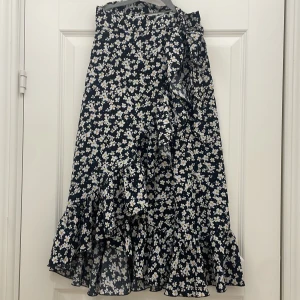 Blommig långkjol med volang - Charmig långkjol med ett blommigt mönster i vitt och gult på en svart bakgrund. Kjolen har en volangdetalj och ett knytband i midjan för extra stil. Perfekt för en somrig look!