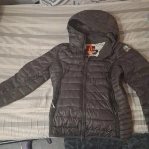 Mörkgrå dunjacka med luva - Säljer en mörkgrå dunjacka från Parajumpers med huva och dragkedja. Jackan har ett quiltat mönster och en broderad logotyp på ärmen. Perfekt för kyliga dagar. Men tyvärr är den inte äkta och har hål under armhålan fast det är svårt att märka 