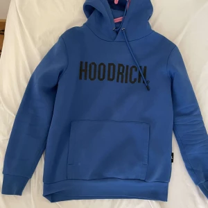 Blå hoodie från Hoodrich - Säljer en snygg blå hoodie från Hoodrich med svart text på bröstet. Den har en klassisk känguruficka och justerbar dragsko i huvan. Perfekt för en avslappnad stil.