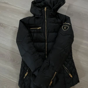 Svart dunjacka från Rockandblue - Snygg svart dunjacka från Rockandblue med huva och flera dragkedjor i guld. Jackan har en quiltad design och är perfekt för kyligare väder. Den har en stilren look med ett märke på ärmen och praktiska fickor med dragkedja. pris kan diskuteras 