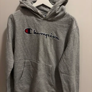 Grå hoodie från Champion - Säljer en klassisk grå hoodie från Champion med deras ikoniska logga på bröstet. Tröjan har en stor ficka framtill och en bekväm huva. Perfekt för en avslappnad stil. Passar som en S 