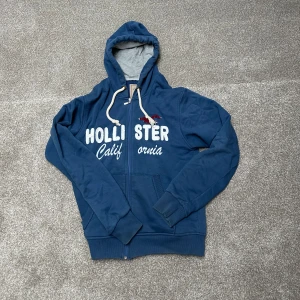 Blå hoodie från Hollister - Säljer en blå hoodie från Hollister med dragkedja och texten 'Hollister California' på framsidan. Den har en justerbar huva med vita snören och långa ärmar. Perfekt för en avslappnad stil.