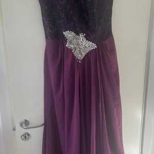 Långklänning i lila med spets och pärlor - Elegant långklänning i mörklila med svart spets upptill och en vacker pärldetalj i midjan. Perfekt för festliga tillfällen med sin glamorösa design. Det finns i Norrköping endast i Norrköping 