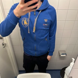 Blå hoodie från Santa Monica Polo Club - Säljer en snygg blå hoodie från Santa Monica Polo Club med dragkedja och tryck på bröstet och ärmen. Den har en justerbar huva med snören och praktiska fickor framtill. Perfekt för en avslappnad stil. Pris går att diskutera. 