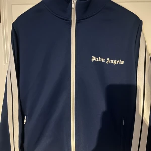 Palm Angels Track Jacket - Säljer denna snygga Palm Angels track jacket i mörkblått med vita detaljer. Jackan är i mycket bra skick, sparsamt använd och utan synliga defekter. Storlek M, passar både herr och dam beroende på önskad passform.  Jag köpte den via Plick och är osäker på om den är äkta, därför säljer jag den till ett lågt pris. Oavsett är det en riktigt stilren och trendig jacka som passar perfekt till vardag. Skicka meddelande vid intresse 