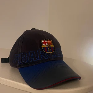 Svart fc Barcelona keps - Säljer en svart keps med broderad FCB-logga på framsidan och texten '1899' baktill. Kepsen har en justerbar rem med klubbens färger och texten 'FCBarcelona' på insidan.
