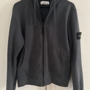 Grå hoodie från Stone Island - Säljer en stilren grå hoodie från Stone Island med dragkedja och praktiska fickor framtill. Hoodien saknar tyvär en knapp på västra fickan som syns på fjärde bilden. Dock är detta inte så märkvärdigt och varan är i perfekt skick annars. Nypris: 4000kr.