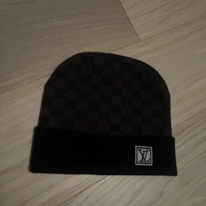 Rutig mössa från Louis Vuitton - Snygg och stilren mössa från Louis Vuitton med ett klassiskt rutigt mönster. Mössan är i en mörk färgton med en uppvikt kant och en diskret logotyp framtill. Perfekt för att hålla värmen med stil.