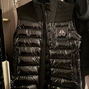 Moose Knuckles väst storlek M - Snygg svart lite glansig dunväst från Moose Knuckles med dragkedja och logotyp på bröstet. Västen har ett quiltat mönster. Köpt från boozt för inte alls längesen. Säljer eftersom jag behöver en storlek mindre. 