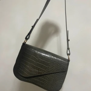 Mörkgrön axelväska med krokodilmönster - Snygg svart axelväska med krokodilmönster. Väskan har en justerbar axelrem och en stilren design som passar perfekt för en trendig look. Perfekt för att bära dina nödvändigheter med stil. Köpt från Nelly.