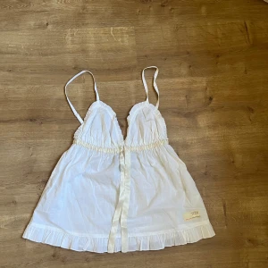Vit tank top med broderi - Säljer en söt vit tank top med smala axelband och volangdetaljer. Toppen har ett fint broderi framtill och knytband i midjan. 
