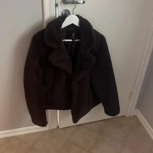 Brun pälsjacka - Säljer en mysig brun pälsjacka med stora slag och knäppning framtill. Jackan har en kortare passform och långa ärmar, perfekt för kyliga dagar. Den är både stilren och bekväm, ett måste i garderoben!
