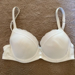 Vit bh - Säljer en elegant vit bh med diskreta ränder från Trofé med spetsdetaljer och en liten pärla framtill. Bh:n har justerbara axelband och en hakknäppning bak. Materialet är en blandning av polyamid och polyester för en bekväm passform.😻Så snygg💋