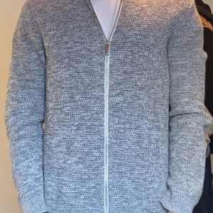 Grå stickad kofta från Selected Homme - Säljer en stilren grå stickad kofta från Selected Homme. Koftan har en dragkedja framtill och är perfekt för kyligare dagar. Den är mjuk och bekväm med långa ärmar och en klassisk passform. Perfekt för att hålla sig varm och snygg!