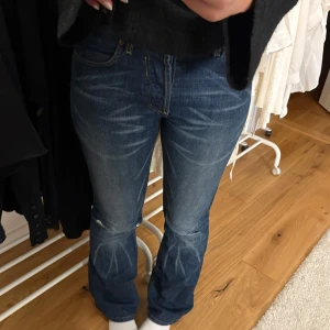 Blå jeans från Lee - Säljer ett par klassiska blå jeans från Lee med vita slitningar. De har en bootcut-stil och är tillverkade i slitstarkt denim. Jeansen sitter under naveln på mig och är perfekta i längden för mig som är 174. Bakfickor med gul kontrastsöm.