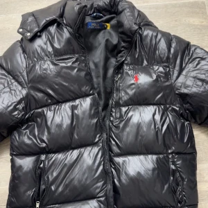 Svart pufferjacka från Polo Ralph Lauren - Säljer en svart pufferjacka från Polo Ralph Lauren. Jackan har en glansig yta med dragkedja och en liten röd logga på bröstet. Perfekt för kyliga dagar med sin vadderade design.