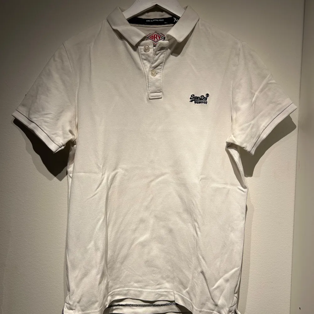 Säljer en stilren vit pikétröja från Superdry. Tröjan har korta ärmar och en klassisk krage med knappar. Perfekt för en avslappnad och snygg look. Superdry-loggan är broderad på bröstet.. T-paidat.