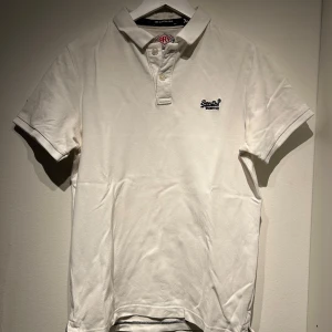 Vit pikétröja från Superdry - Säljer en stilren vit pikétröja från Superdry. Tröjan har korta ärmar och en klassisk krage med knappar. Perfekt för en avslappnad och snygg look. Superdry-loggan är broderad på bröstet.