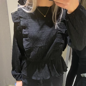Svart blus med volangdetaljer - Snygg svart blus med volangdetaljer längs framsidan och ärmarna. Blusen har en smickrande passform och är perfekt för att ge en stilfull touch till din outfit. Den är tillverkad i ett texturerat material som ger en unik look.