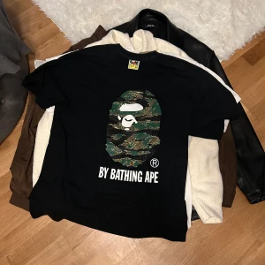 Svart t-shirt från A Bathing Ape - Säljer en svart t-shirt från A Bathing Ape med det ikoniska kamouflagemönstret och logotypen på framsidan. T-shirten har en normal passform och är kortärmad. Perfekt för streetwear-entusiaster.