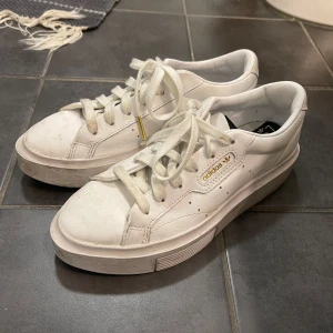 Vita sneakers från Adidas - Säljer ett par vita sneakers från Adidas med snörning och en klassisk design. Skorna har en tjock sula och guldiga detaljer vid snörningen. Perfekta för en stilren look.