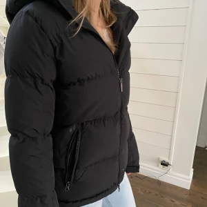 Svart dunjacka från Kingsland - Snygg svart dunjacka från Kingsland med huva och dragkedja. Perfekt för kyliga dagar. Jackan har en stilren design med logotyp på bröstet och praktiska fickor med dragkedja.