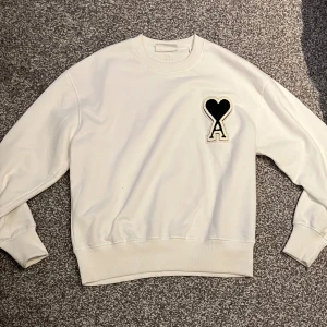 Vit sweatshirt från Ami Alexandre Mattiussi - Säljer en stilren vit sweatshirt från Ami Alexandre Mattiussi med ett unikt hjärta och bokstaven A broderat på bröstet. Tröjan har långa ärmar och en bekväm passform. Perfekt för en avslappnad look.