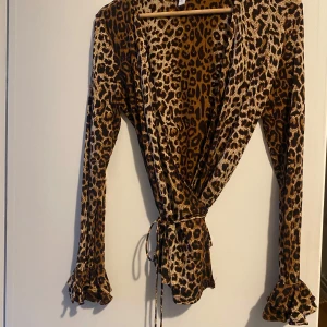 Leopardmönstrad omlottblus från Other stories  - Snygg leopardmönstrad omlottblus från other stories med långa ärmar och volangdetaljer vid ärmsluten. Blusen har en djup v-ringning och knytband i midjan för en justerbar passform. Perfekt för att ge en trendig touch till din outfit 🐆