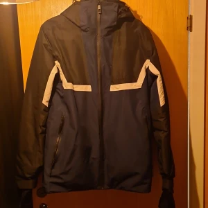 Blå och svart jacka med huva - Säljer en blå och svart jacka med huva och dragkedja. Jackan har vita detaljer på ärmarna och bröstet. Perfekt för kyligare dagar.