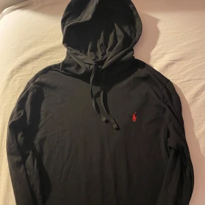Svart hoodie från Ralph Lauren - Säljer en stilren svart hoodie från Ralph Lauren med det klassiska röda broderade logotypen på bröstet. Tröjan har en justerbar huva med snören och långa ärmar. Perfekt för en avslappnad look. Tyget är ganska tunt.