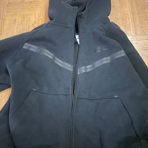 Svart hoodie från Nike - Säljer en svart hoodie från Nike med dragkedja och huva. Den har diskreta ränder över bröstet och sidofickor. Perfekt för en avslappnad stil.          Skulle säga ganska liten för L så mer som en M