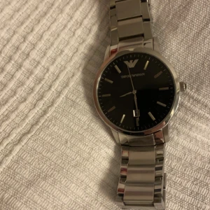 Silverfärgad klocka från Emporio Armani - Snygg och stilren klocka från Emporio Armani i silverfärgat rostfritt stål. Den har en svart urtavla med minimalistiska markeringar och datumvisning. Perfekt för den som gillar en klassisk och elegant stil.
