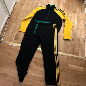 Svart och gul träningsdräkt från Adidas - Snygg svart och gul träningsdräkt från Adidas med dragkedja framtill och gröna detaljer. Dräkten har långa ärmar och byxor med gula ränder längs sidorna. Perfekt för träning eller en sportig look.