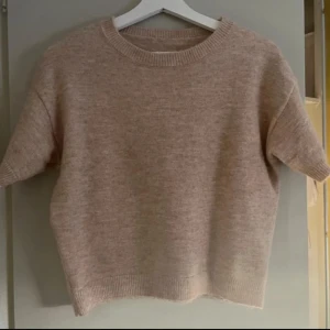 Beige stickad t-shirt - Säljer en mysig beige stickad tröja med korta ärmar. Perfekt för lager-på-lager under kyligare dagar. Tröjan har en rund halsringning och ribbade kanter för en stilren look. Passar utmärkt till både jeans och kjol.