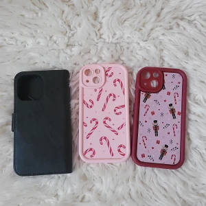 Tre mobilskal för iPhone 15 - Säljer tre olika mobilskal. Ett svart skinnfodral, ett rosa skal med polkagrisar och ett lila skal med julmotiv. Perfekt för att ge din mobil en ny look!
