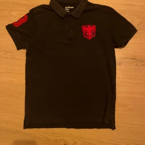 Svart pikétröja med röd broderad detalj - Snygg svart pikétröja från Giorgio Di Mare med en röd broderad örn på bröstet. Tröjan har en klassisk krage med röd och vit randig insida och knappar framtill. Perfekt för en stilren look.