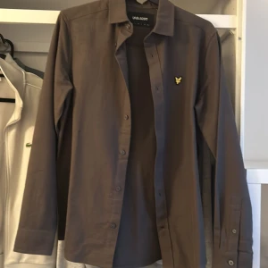Brun skjorta från Lyle & Scott - Snygg brun skjorta från Lyle & Scott med klassisk krage och knappar framtill. Skjortan har ett broderat emblem på bröstet som ger en stilren touch. Perfekt för en avslappnad men ändå uppklädd look.