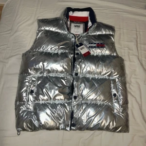 Tommy Hilfiger väst - Den här västen är helt ny (oanvänd), det finns inga repor eller skador på västen. Silverfärgad, zip fickor, puffig, riktigt varm, storlek L. Tag är med. Det går att justera midjan också. Mitt pris är 1050kr, nypriset är 2200kr. Kan inte ladda mer än 5 bilder, men kan skicka flera bilder privat om det behövs. 
