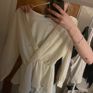 Vit omlottblus - Säljer en stilren vit omlottblus. Blusen har långa ärmar och en v-ringad design med knytning i midjan. Perfekt för en elegant look.