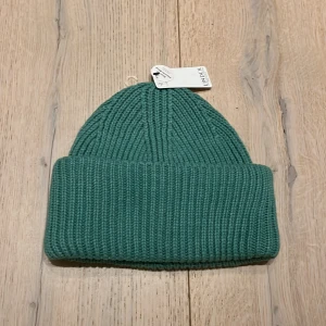 Grön stickad mössa från Lindex - Snygg grön stickad mössa från Lindex i återvunnen polyesterblandning. Perfekt för kyliga dagar med sin ribbade design som ger en skön passform. En stilren accessoar för både höst och vinter.