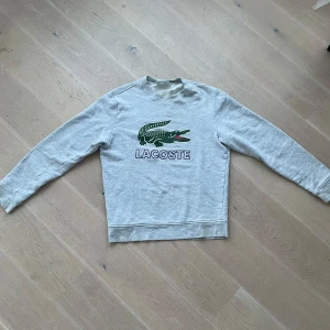 Grå sweatshirt från Lacoste - Säljer en stilren grå sweatshirt från Lacoste med det ikoniska krokodilmärket på bröstet. Tröjan har långa ärmar och en klassisk rund halsringning. Perfekt för en avslappnad look.