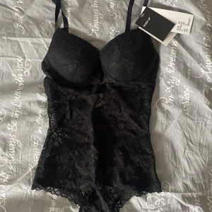 Svart spetsbody från H&M - Säljer en elegant svart spetsbody från H&M. Den har justerbara axelband och en vacker blommig spetsdesign. Perfekt för att ge en lyxig känsla till din outfit. Ny med taggar kvar.