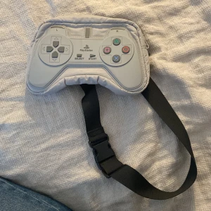 Grå axel/mag-väska i form av PlayStation-kontroll från Zara - Säljer en unik grå maglväska från Zara, designad som en PlayStation-kontroll. Väskan har en justerbar svart rem och stängs med dragkedja. Perfekt för gaming-entusiaster som vill ha något annorlunda!