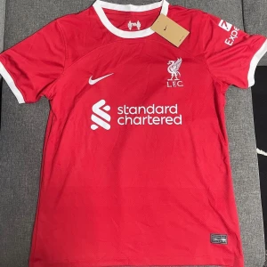 Röd Liverpool fotbollströja - Säljer en röd Liverpool fotbollströja från Nike. Den har korta ärmar och är i nyskick. Tröjan har vita detaljer och klubbens logga på bröstet. Perfekt för alla Liverpool-fans!