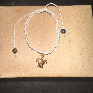 Vitt armband med sköldpaddshänge - Säljer ett superfint vitt armband med ett gulligt sköldpaddshänge i guld. Armbandet har justerbara snören med svarta pärlor i ändarna. Perfekt för att ge en strandig vibe till din outfit!