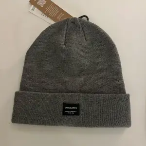 Säljer en stilren grå mössa från Jack & Jones. Den är i en klassisk design med uppvikt kant och en liten svart etikett med märkesloggan framtill. Perfekt för att hålla sig varm under kyliga dagar.