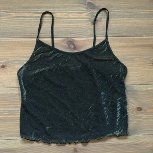 Svart glittrig linnetopp från First Position - Säljer en snygg svart glittrig see through linnetopp från First Position. Toppen har tunna axelband och är perfekt för festliga tillfällen. Materialet är mjukt och skimrar i ljuset, vilket ger en extra touch av glamour.