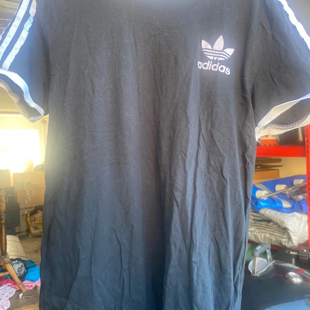 Säljer en svart t-shirt från Adidas med klassiska vita ränder längs ärmarna. T-shirten har en normal passform och är kortärmad. Perfekt för en avslappnad stil eller träning. Adidas-loggan syns tydligt på bröstet. Skön och stilren design!. T-shirts.